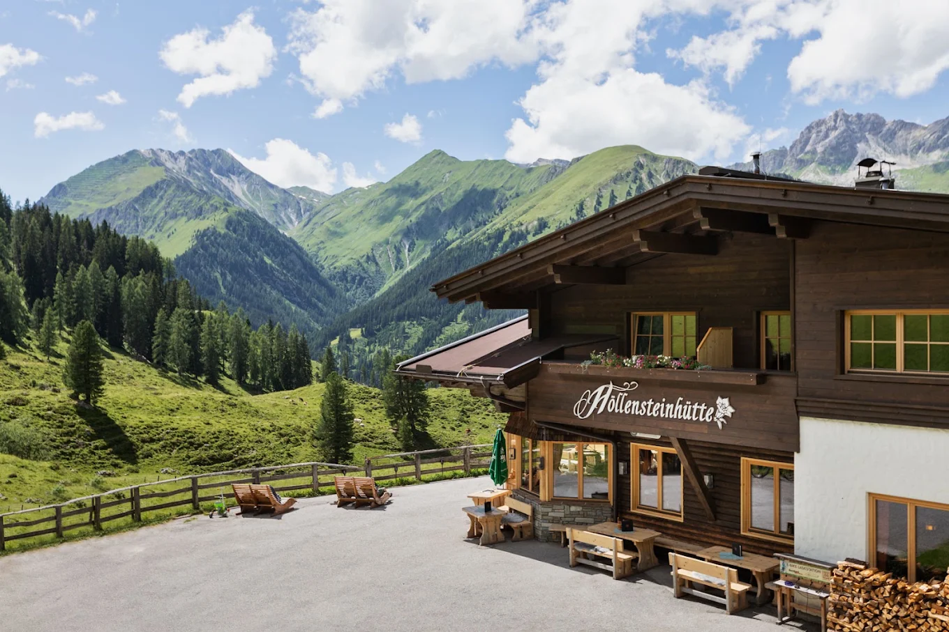 The Höllensteinhütte, a dark-timbered mountain inn with flower boxes and wooden benches, looking west down the green Tux valley towards the Tuxer Alps.