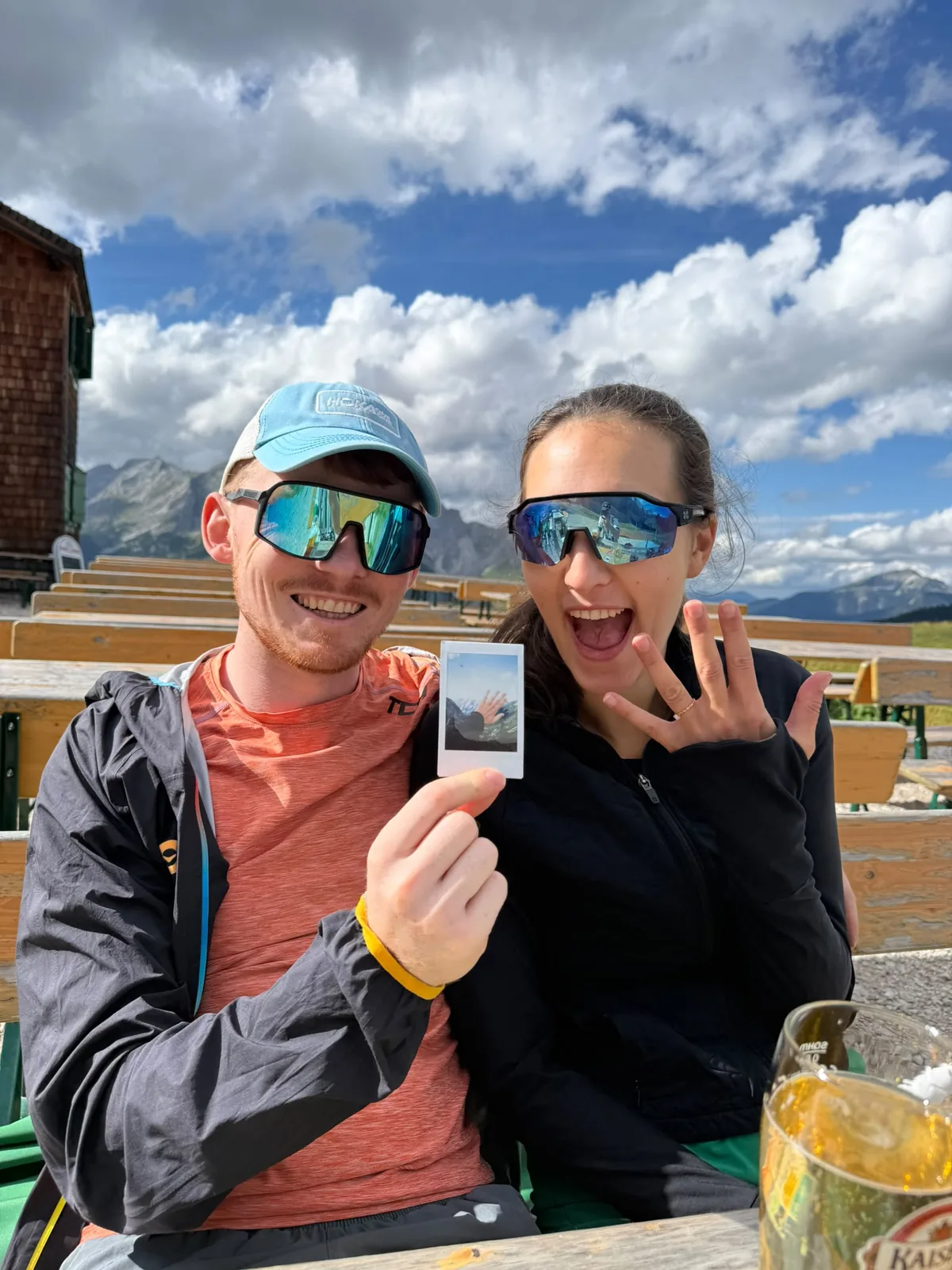 Hannah and Angus at a mountain hut terrace, holding a polaroid, with a beer on the table and benches behind.